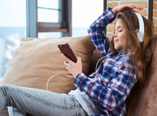 Beautiful little girl in headphones using smartphone on a couch - 写真・画像