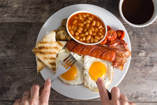 Mulher comendo um café da manhã tradicional inglês com ovos fritos, salsichas, feijão, cogumelos, tomates grelhados, bacon, café e suco de laranja, na mesa de madeira
 - Foto, Imagem