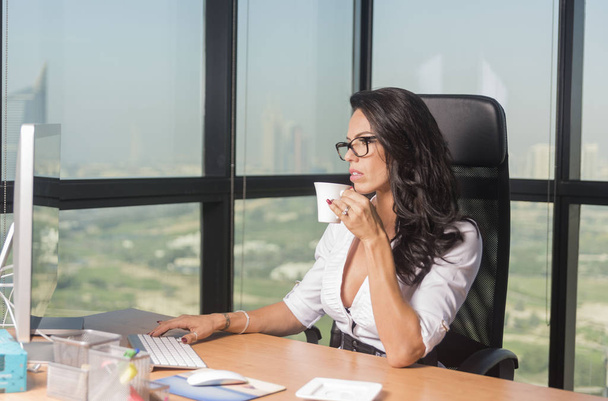 Kaunis sihteeri tai liiketoiminnan nainen toimistossa lasit siemaillen teetä tietokoneella
 - Valokuva, kuva