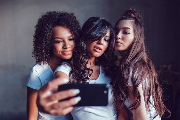 Three beautyful smiling girls in white t-shirt grimace and make selfie using smartphone. - Photo, Image