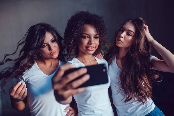 Three beautyful smiling girls in white t-shirt grimace and make selfie using smartphone. - Photo, Image