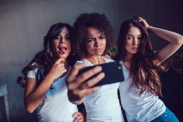 Trzy beautyful uśmiechniętych dziewcząt w biały t-shirt grymas i zrobić autoportret za pomocą smartfona. - Zdjęcie, obraz
