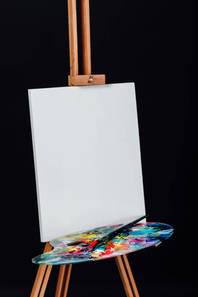 Tools of the artist. Brushes, wooden easel tripod, palette colorful. Black background, studio, nobody. - Photo, image