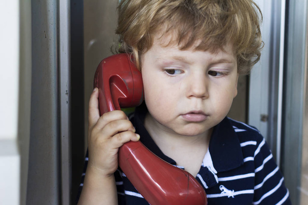 O bebé a falar ao telefone fixo. Um rapaz numa cabine telefónica. Aparelho vermelho
 - Foto, Imagem