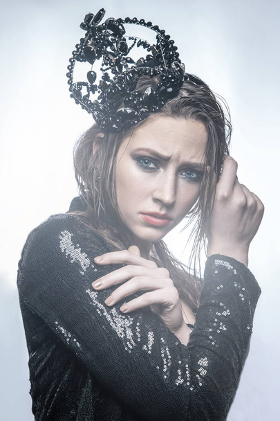 fashion beauty portrait of young beautiful young woman with makeup and freckles on her face with black crown on her head and black dress. with light from behind on light background. - 写真・画像