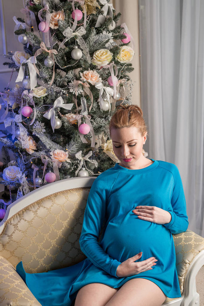 Pregnant girl in a blue tunic sitting in a chair next to the Christmas tree - Foto, Imagen