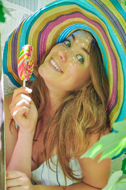funny girl wearing a colorful hat with lollipop in window with grape leaves - Photo, image