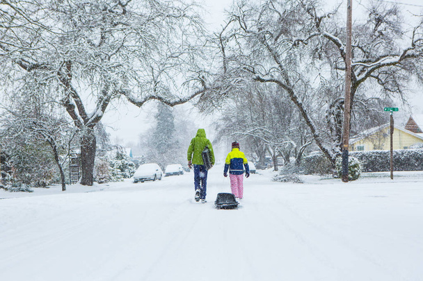 Τα παιδιά με τα πόδια κάτω από το κέντρο του ένα άδειο δρόμο σέρνει έλκηθρα στο χιόνι φρέσκο πεσμένο χειμώνα. - Φωτογραφία, εικόνα