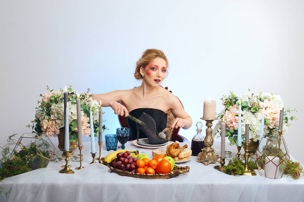 Fashion blonde model with colorful makeup sitting behind festive decorated table and cutting suede shoe - Fotografie, Obrázek