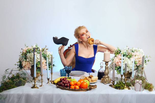 Fashion blonde model with colorful makeup sitting behind festive decorated table and eating felt shoe - Фото, зображення