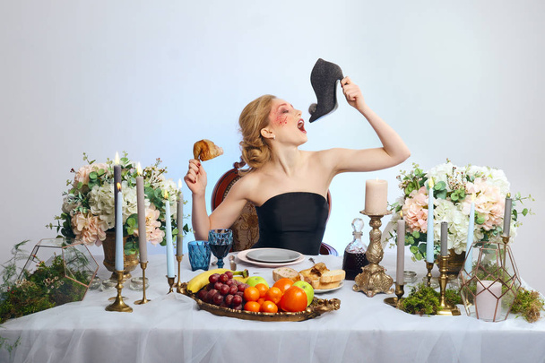 Fashion blonde model with colorful makeup sitting behind festive decorated table and eating felt shoe - Foto, immagini