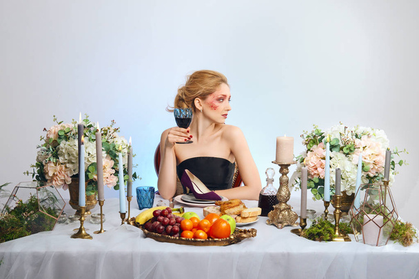 Fashion blonde model with colorful makeup drinking wine behind decorated table and eating suede shoe - Фото, зображення