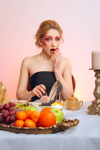 Tricky lady with colorful makeup sitting behind festive table and eating felt shoe - Photo, image