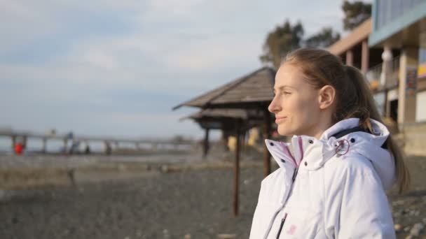 Młoda kobieta w białej kurtce w zimie na plaży o zachodzie słońca - Materiał filmowy, wideo