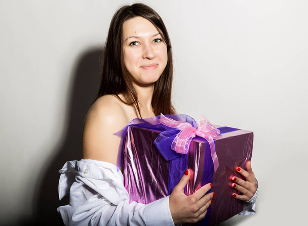Portrait of young beautiful smiling girl in a mans white shirt, holding gift and enjoys - Photo, Image