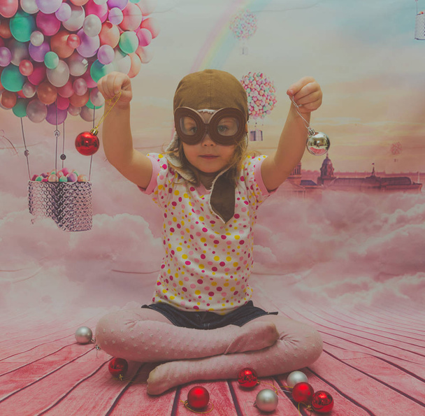 the girl in the pilot's helmet and glasses plays with red Christmas balls - Fotoğraf, Görsel