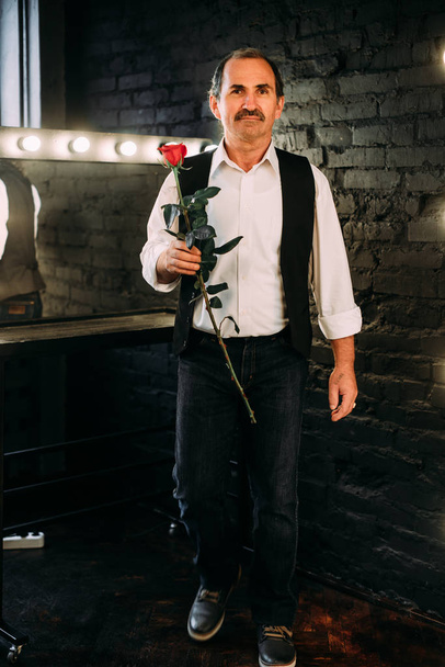 中年男性は白いシャツと黒いベストに身を包んだ赤いバラの花を保持しているハンサムな中間独身。黒の背景. - 写真・画像