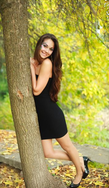 Pretty smiling girl standing under a tree in the autumn park - Zdjęcie, obraz