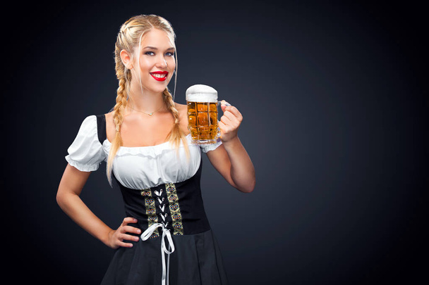 Young sexy Oktoberfest waitress, wearing a traditional Bavarian dress, serving big beer mugs on black background. - Foto, afbeelding