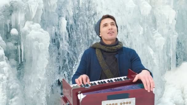Happy young man playing the harmonium on a frozen waterfall - Footage, Video