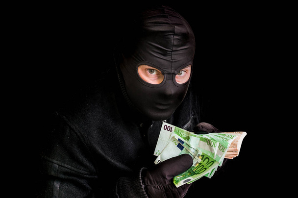 Masked thief in balaclava with stolen money isolated on black - Photo, Image
