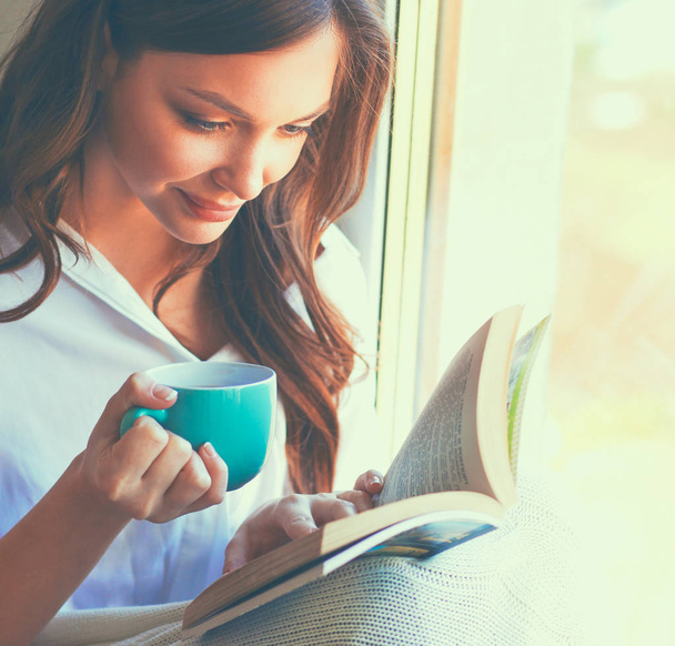 Young woman at home sitting near window relaxing in her living room reading book and drinking coffee or tea - Photo, image