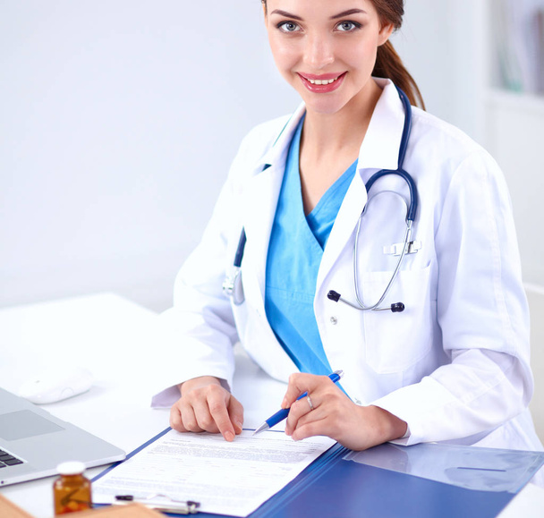 Bonito jovem sorridente médico feminino sentado na mesa e escrevendo. - Foto, Imagem