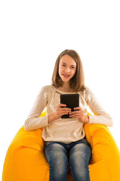Young smiling girl holding the tablet while sitting on yellow be - Фото, изображение