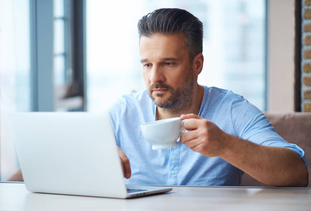 Hombre guapo con taza de café usando el ordenador portátil en casa
 - Foto, Imagen