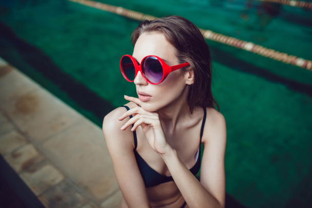Belle femme brune en lunettes de soleil drôles avec un maquillage professionnel assis près de la piscine
. - Photo, image