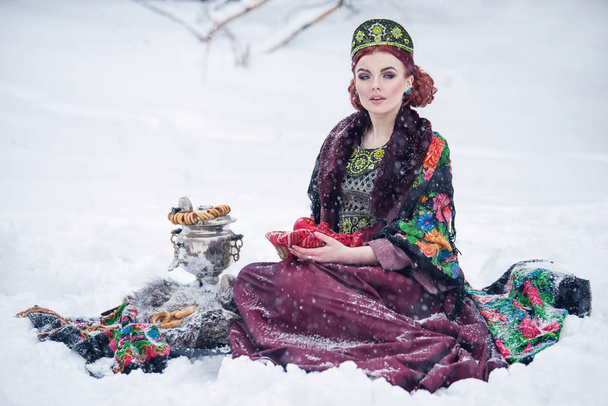 Portrait d'une magnifique jeune femme en robe de style russe lors d'une forte gelée par une journée enneigée d'hiver avec des pommes et du samovar. Fille modèle russe en coiffure traditionnelle
. - Photo, image