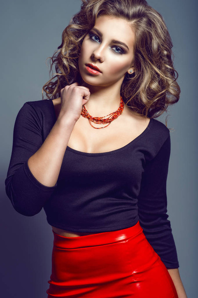 Porträt der jungen hübschen Model mit langen welligen Haaren trägt schwarzes Top mit drei Viertel Ärmel und Bootsausschnitt und rotem Lederrock mit roten Perlen am Hals - Foto, Bild