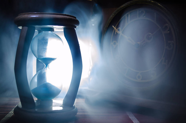 Концепция времени Силуэт часов Песочные часы и старые винтажные деревянные часы со стрелками и дымом на темном фоне с горячим желтым оранжевым синим холодным задним освещением, или символы времени с копировальным пространством
 - Фото, изображение