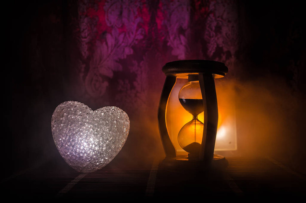 Valentine's Day composition with sweet burning multicolored heart on dark background and Silhouette of Hourglass clock, time and love concept, selective focus, or love greeting card of heart red green - Photo, Image