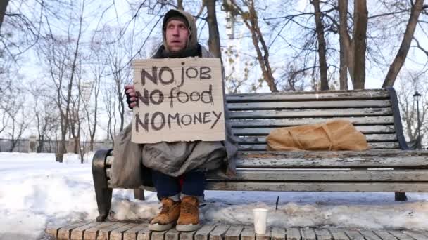  4 k. 失業者。段ボールを持つ男は、冬の公園でベンチに座っています。ドーリー ショット  - 映像、動画