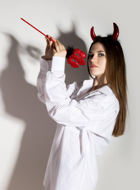 jong meisje in een witte overhemd met rode hoorns houden van trident mans en ziet eruit als mooie duivel - Foto, afbeelding