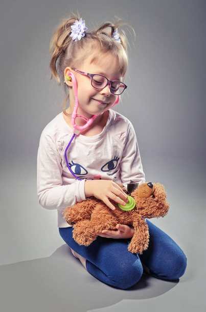 Menina bonita joga no médico trata um ursinho de pelúcia em uma ba cinza
 - Foto, Imagem