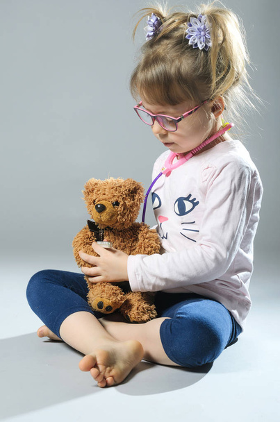 Mooi meisje speelt in de arts behandelt een teddybeer op een grijze ba - Foto, afbeelding