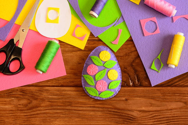 Пасхальное яйцо украшают цветы, ножницы, нитки, игла, бумажный узор, цветные войлочные листы и объедки на деревянном столе. Пасхальный подарок ручной работы для детей. Вид сверху
 - Фото, изображение