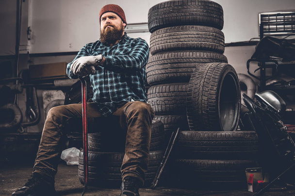 Parrakas mekaanikko istuu vanhojen renkaiden päällä
 - Valokuva, kuva
