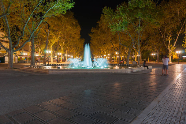 beleuchteter Brunnen und baumgesäumte Promenade - Foto, Bild