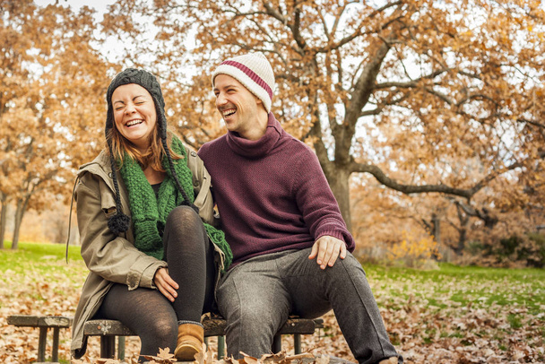 Young adult couple laughing, sit in a bench. Park full of leaves - Photo, Image