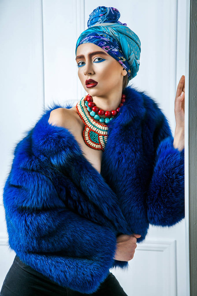 Beauty portrait of fashion model with colored headwear, blue fur coat red eyebrow and lips makeup and necklace. studio shot near windows and white wall - Fotoğraf, Görsel