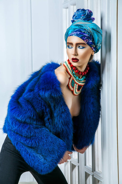 Beauty portrait of fashion model with colored headwear, blue fur coat red eyebrow and lips makeup and necklace. studio shot near windows and white wall - Foto, afbeelding
