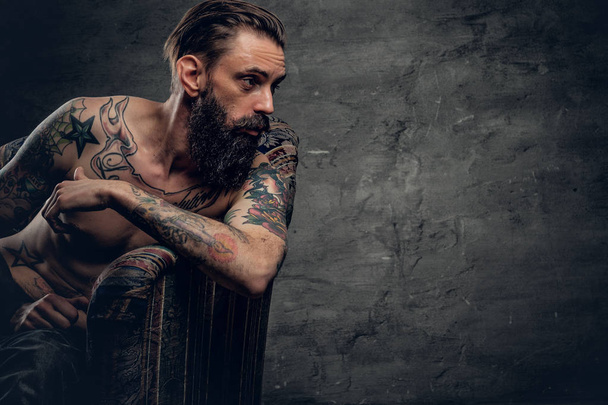 Без сорочки, татуйований чоловік сидить на стільці
 - Фото, зображення