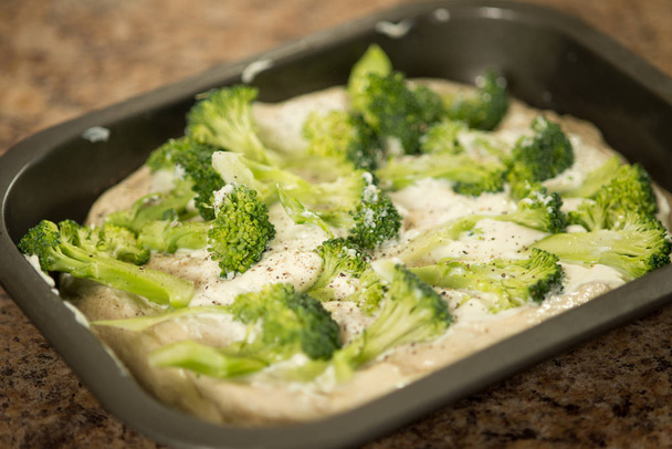 Rauw brooddeeg in kleine bakplaat, afgevlakt en gegarneerd met ongekookte broccoli - Foto, afbeelding
