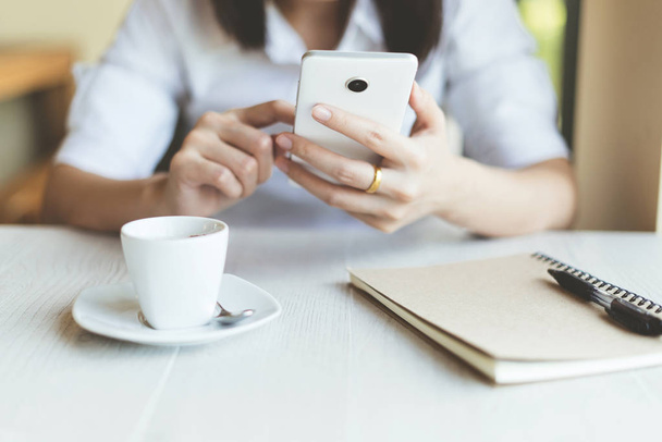 Νεαρή γυναίκα χρησιμοποιώντας smartphone με ένα φλιτζάνι καφέ και το Σημειωματάριο. - Φωτογραφία, εικόνα