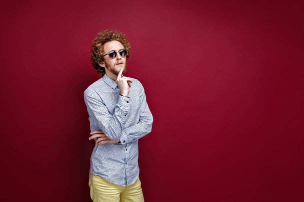 Stylish curly-haired man in sunglasses on red background. Thinki - Photo, image