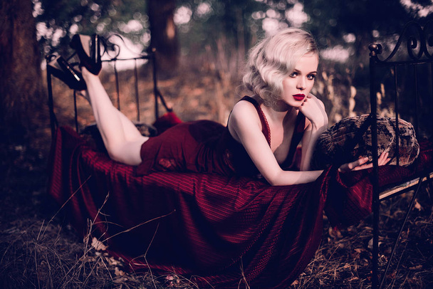 Beautiful and elegant blonde woman with red lips and hair waves wearing wine red nightie posing on the bed outdoors autumn, retro vintage style and fashion. Retouched toning shot - Photo, Image
