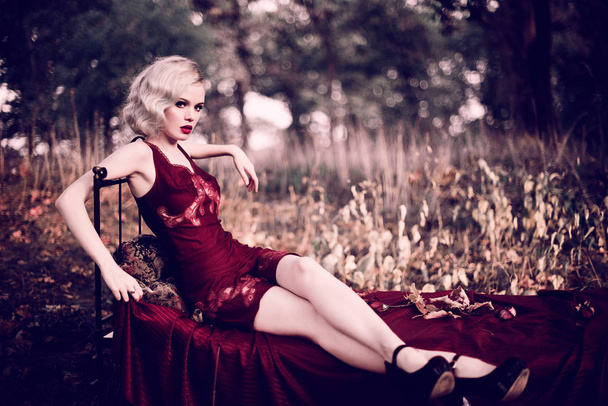 schöne und elegante blonde Frau mit roten Lippen und Haaren Wellen tragen weinroten Nachthemd posiert auf dem Bett im Freien Herbst, Retro-Vintage-Stil und Mode. retuschierte Tonung Schuss - Foto, Bild